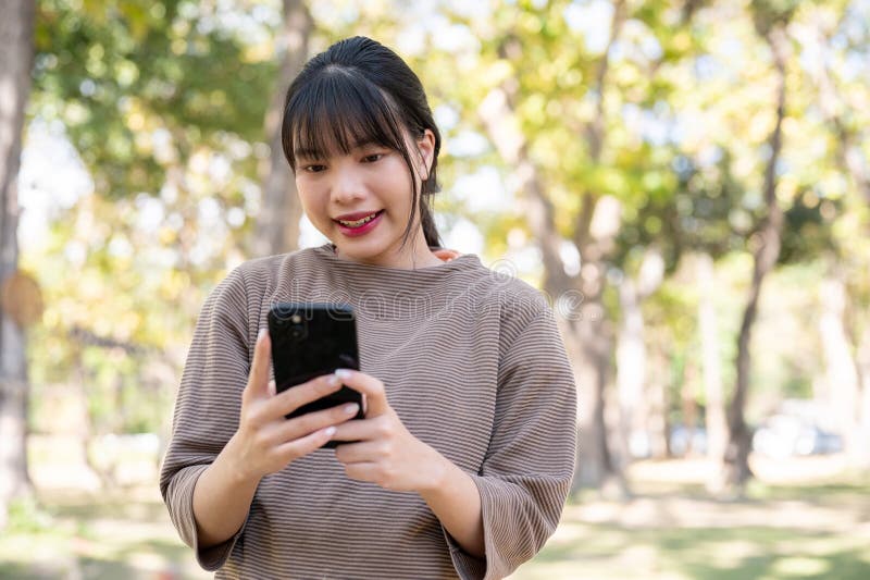 一位美丽的亚洲女子在公园散步时，正用智能手机和朋友聊天 免版税库存照片