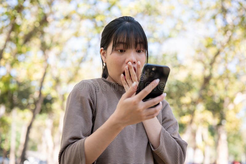 一位惊讶的亚洲女性正在公园里散步时，在她的智能手机上阅读意外的好消息 库存照片