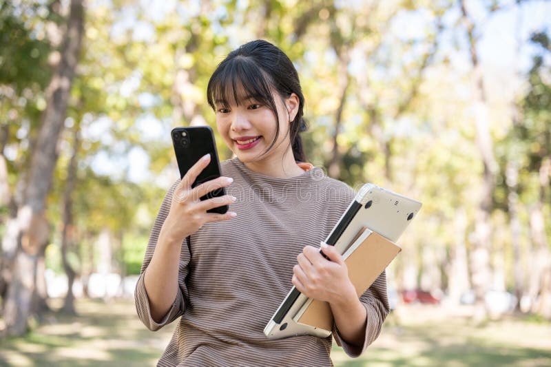一位年轻漂亮的亚洲女学生正在校园公园里使用她的智能手机 库存图片