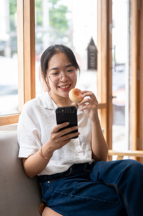在咖啡馆的沙发上用智能手机享受甜甜圈的快乐的亚洲年轻女性 免版税库存图片