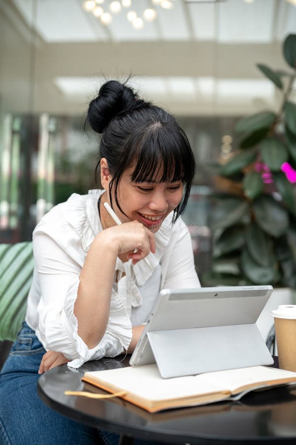 年轻快乐的亚洲女性正享受着在咖啡馆里阅读其数字平板电脑上的东西 免版税库存照片