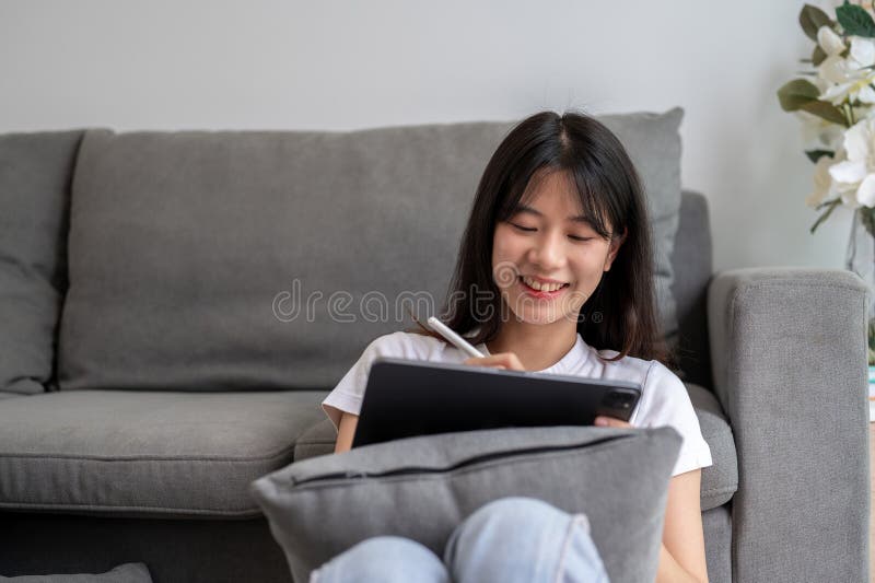 在客厅的数字平板上用手写笔的年轻幸福亚洲女性 图库摄影