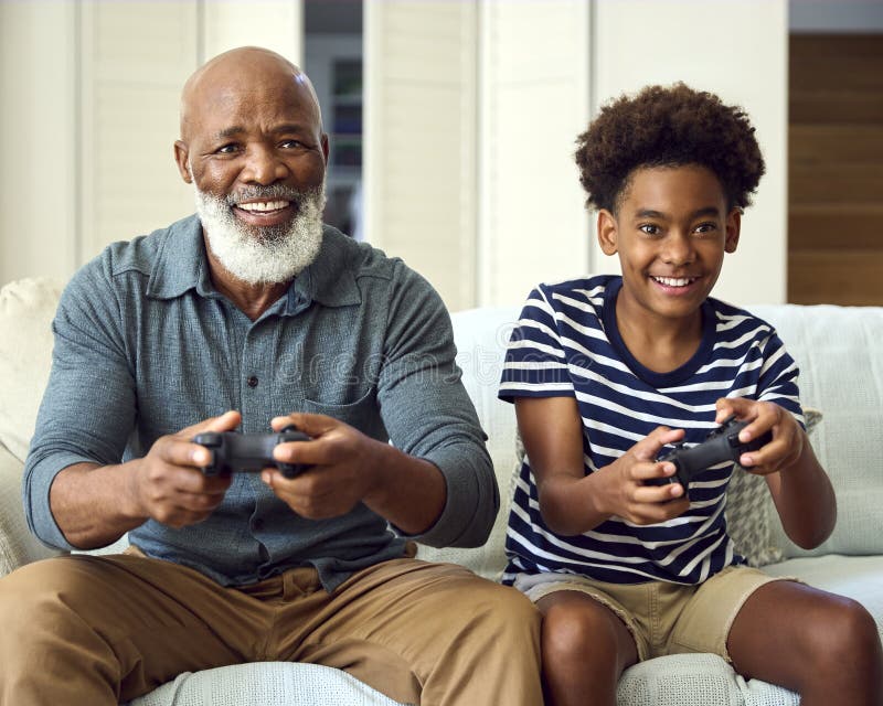 Nonno e nipote sul divano a casa giocano a videogioco insieme a un ragazzo che festeggia fotografie stock