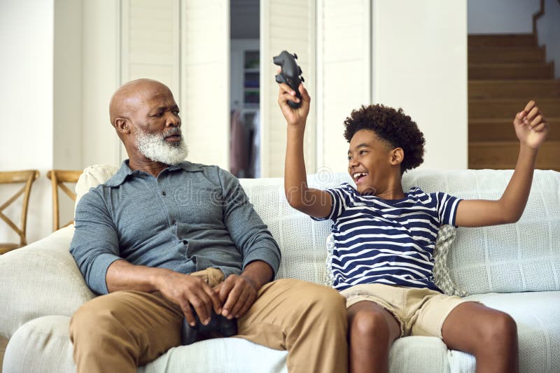 Nonno e nipote sul divano a casa giocano a videogioco insieme a un ragazzo che festeggia immagini stock libere da diritti