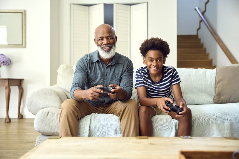 Nonno e nipote sul divano a casa giocano a videogioco insieme a un ragazzo che festeggia fotografie stock