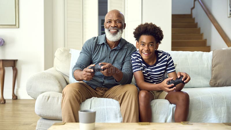 Nonno e nipote sul divano a casa giocano a videogioco insieme a un ragazzo che festeggia fotografia stock
