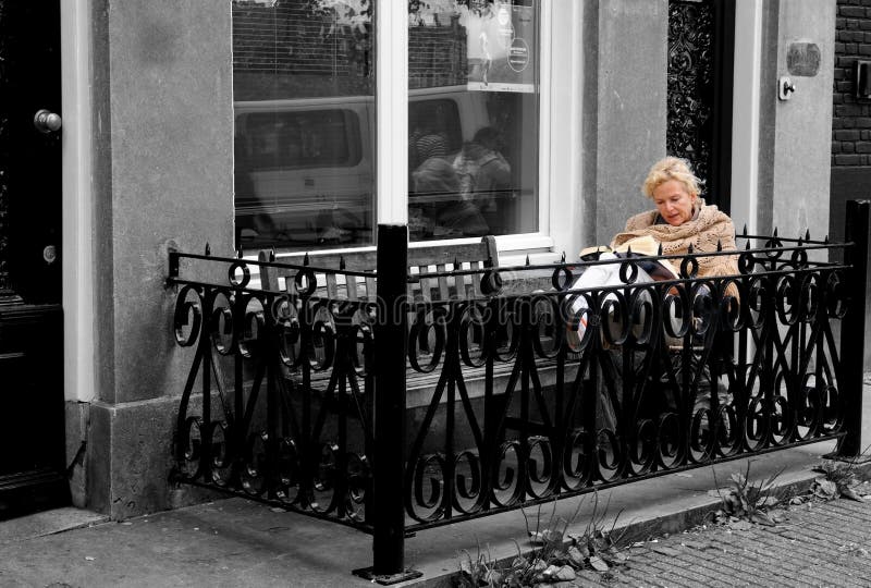 Amsterdam, 2010 - Beautiful elder blonde woman bundled up and reading a book on a balcony at street level. Image background in Black and white. Amsterdam, 2010 - Beautiful elder blonde woman bundled up and reading a book on a balcony at street level. Image background in Black and white.