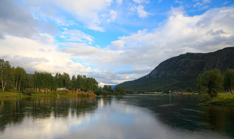 Breathtaking Norwegian lake landscape at sunset with dramatic cloud formations reflected in calm waters. The peaceful scene showcases a pristine lake surrounded by birch and evergreen forests, with majestic mountains rising in the background. Golden evening light illuminates the clouds while their perfect mirror reflection doubles the visual impact. Small houses dot the shoreline, nestled among the trees, representing the harmonious coexistence of human settlement with pristine Nordic nature. This tranquil Scandinavian vista captures the essence of Norway's natural beauty with its combination of water, mountains, and forests. The serene atmosphere is enhanced by the still water surface that acts as a natural mirror, creating stunning symmetry in the composition. Perfect stock image for travel destinations, nature conservation, eco-tourism, or Scandinavian lifestyle concepts. The warm sunset colors contrast beautifully with the cool blues of the water and the deep greens of the forested hillsides. This idyllic scene represents the untouched wilderness areas that make Norway famous for its spectacular landscapes. The image conveys themes of tranquility, natural beauty, and environmental preservation. Ideal for promoting outdoor activities, meditation retreats, or showcasing the scenic wonders of Northern Europe. The dramatic sky with its mix of white and gray clouds adds depth and visual interest to this stunning natural panorama of a typical Norwegian lake surrounded by mountains and forests. Stock image photography. Contrast enhanced stock images, royalty-free photos and pictures