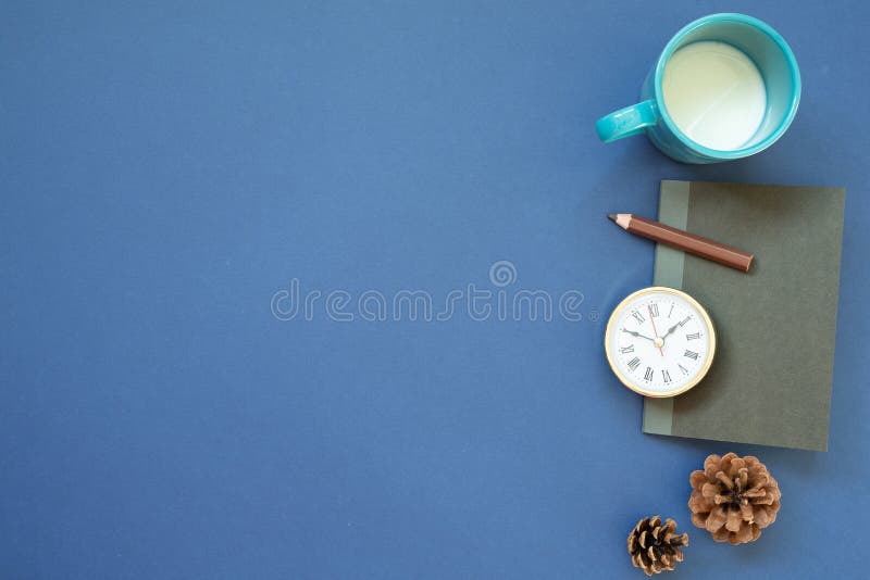 Notebook, clock, colored pencil, cup of milk with pine cone on navy blue background. top view, copy space. workspace. studio shot