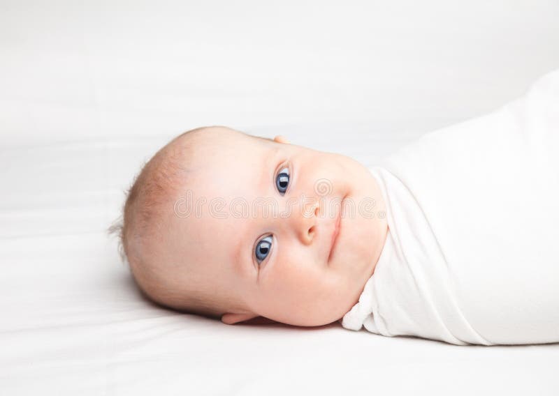 Close-up shot of three month baby girl swaddled in white blanket laying on a bed. Swaddling is a practice of wrapping infants in cloths in order to prevent limb movement. Medical and psychological effects of swaddling are controversial. Close-up shot of three month baby girl swaddled in white blanket laying on a bed. Swaddling is a practice of wrapping infants in cloths in order to prevent limb movement. Medical and psychological effects of swaddling are controversial.