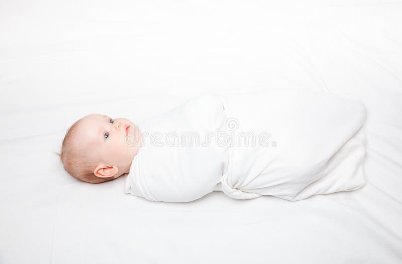 Three month baby girl swaddled in white blanket laying on a bed. Swaddling is a practice of wrapping infants in cloths in order to prevent limb movement. Medical and psychological effects of swaddling are controversial. Three month baby girl swaddled in white blanket laying on a bed. Swaddling is a practice of wrapping infants in cloths in order to prevent limb movement. Medical and psychological effects of swaddling are controversial.