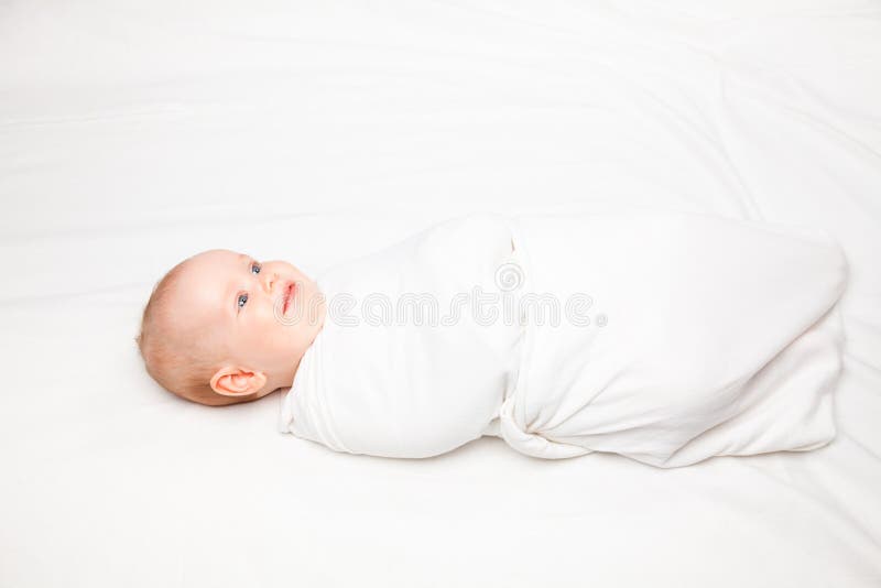 Three month baby girl swaddled in white blanket laying on a bed. Swaddling is a practice of wrapping infants in cloths in order to prevent limb movement. Three month baby girl swaddled in white blanket laying on a bed. Swaddling is a practice of wrapping infants in cloths in order to prevent limb movement