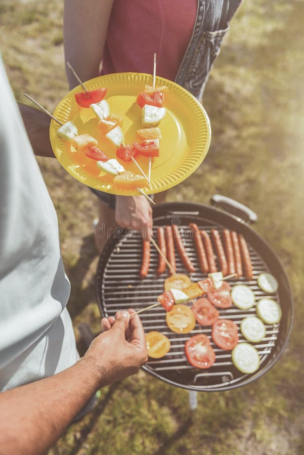 Male hands holding plastic plate with vegetable skewers on it. Man and woman standing near brazier in nature. Top view close up. Male hands holding plastic plate with vegetable skewers on it. Man and woman standing near brazier in nature. Top view close up