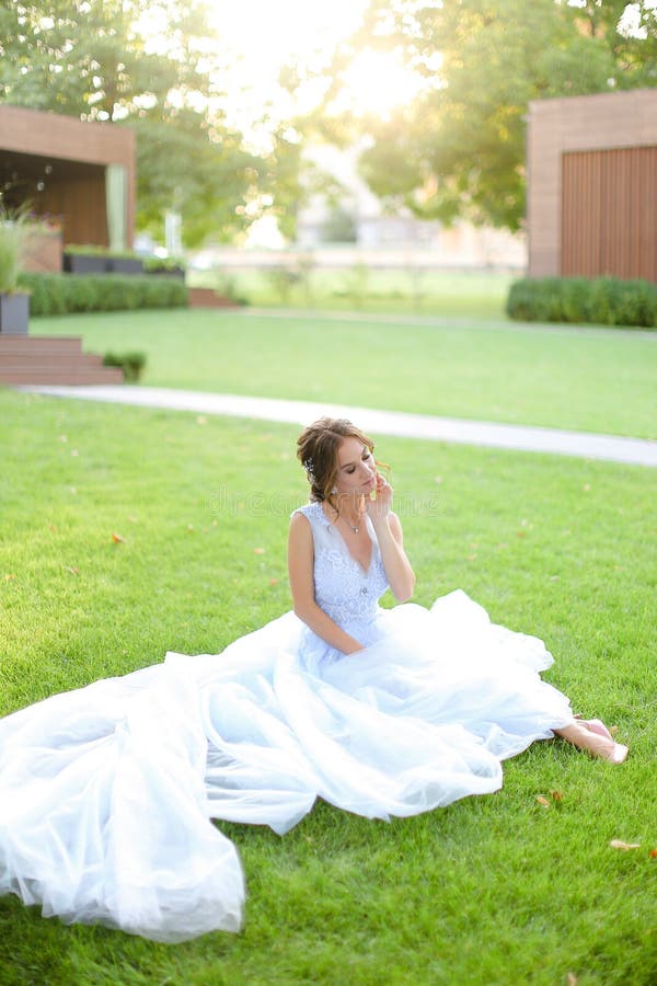 Novia caucásica que se sienta en hierba en el parque, vestido blanco que lleva fotografía de archivo Novia caucásica que se sienta en hierba en el parque, vestido blanco que lleva fotografía de archivo