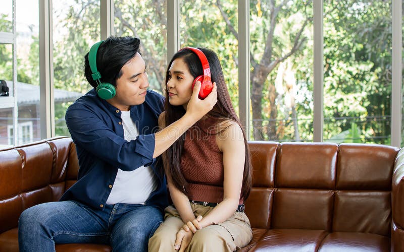 Novia de pareja de amigos asiáticos sentada en un sofá en una acogedora sala de estar en casa por la mañana usando auriculares par fotografía de archivo