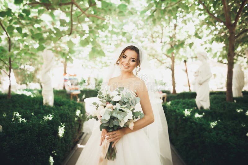 Novia morena hermosa en el vestido blanco elegante que sostiene el ramo que presenta árboles aseados fotografía de archivo Novia morena hermosa en el vestido blanco elegante que sostiene el ramo que presenta árboles aseados fotografía de archivo