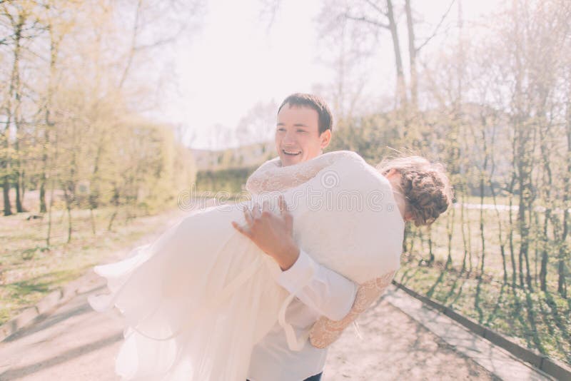 Novio de risa hermoso que lleva a la novia hermosa en sus brazos en el parque de la primavera imágenes de archivo libres de regalías Novio de risa hermoso que lleva a la novia hermosa en sus brazos en el parque de la primavera imágenes de archivo libres de regalías