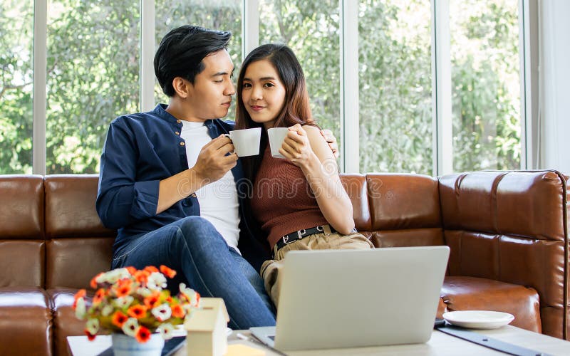 Novio y novia de una pareja asiática sentada en un sofá en una acogedora sala de estar en casa por la mañana tomando café juntos p foto de archivo