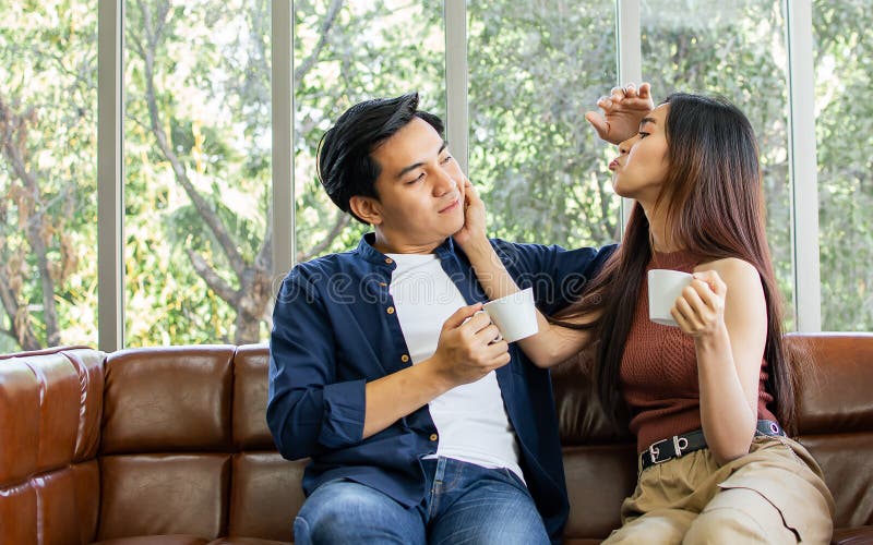 Novio y novia de una pareja asiática sentada en un sofá en una acogedora sala de estar en casa por la mañana tomando café juntos p imagen de archivo