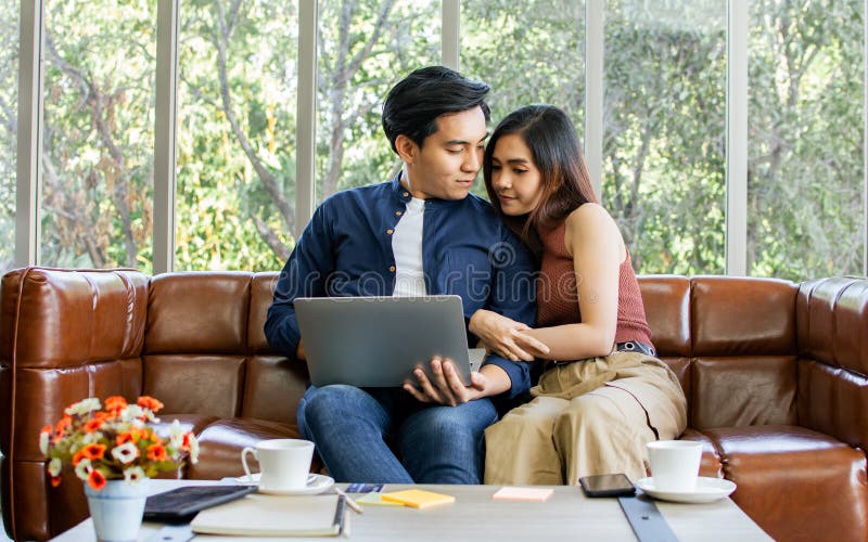 Novio y novia de una pareja asiática sentada en un sofá en una acogedora sala de estar en casa por la mañana usando una laptop par imagen de archivo libre de regalías