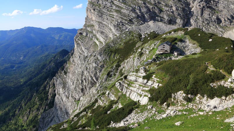 Nowa alpejska chata na zielonej trawie otoczona potężnymi skałami z zalesionym grzbietem górskim w tle obrazy royalty free Nowa alpejska chata na zielonej trawie otoczona potężnymi skałami z zalesionym grzbietem górskim w tle obrazy royalty free