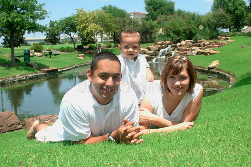 Young married couple with their toddler child. New family taking pictures in white shirts and tan shorts. Family of three. Family laying on the ground with park behind them. Young married couple with their toddler child. New family taking pictures in white shirts and tan shorts. Family of three. Family laying on the ground with park behind them.