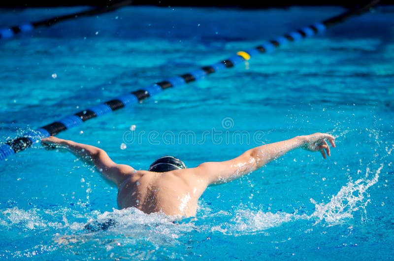 Young man swimming butterfly in an outdoor pool, seen from behind, during a competition. Black and blue lane lines stretch diagonally across the frame, moving into shadow at the edge of the frame. Arms are outstretched, with water drops spraying off. Water is blue, splashes bright white. Sunny summer day. Young man swimming butterfly in an outdoor pool, seen from behind, during a competition. Black and blue lane lines stretch diagonally across the frame, moving into shadow at the edge of the frame. Arms are outstretched, with water drops spraying off. Water is blue, splashes bright white. Sunny summer day.