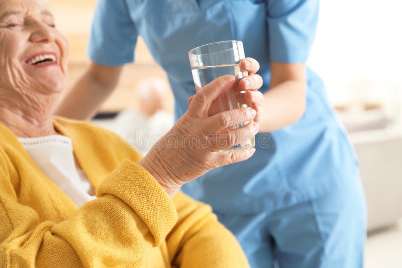 Nurse giving glass of water to senior woman indoors. Assisting elderly people