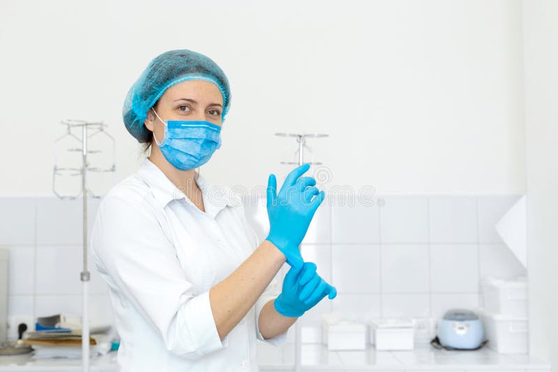 A nurse in a white coat puts on rubber gloves before a medical procedure in a bright handling room.