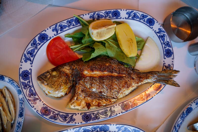 Ny grillad hel fisk för havsbraxen med den grillade potatisen, grön sallad, tomaten och gul limefrukt på den vita ovala plattan m royaltyfria bilder Ny grillad hel fisk för havsbraxen med den grillade potatisen, grön sallad, tomaten och gul limefrukt på den vita ovala plattan m royaltyfria bilder