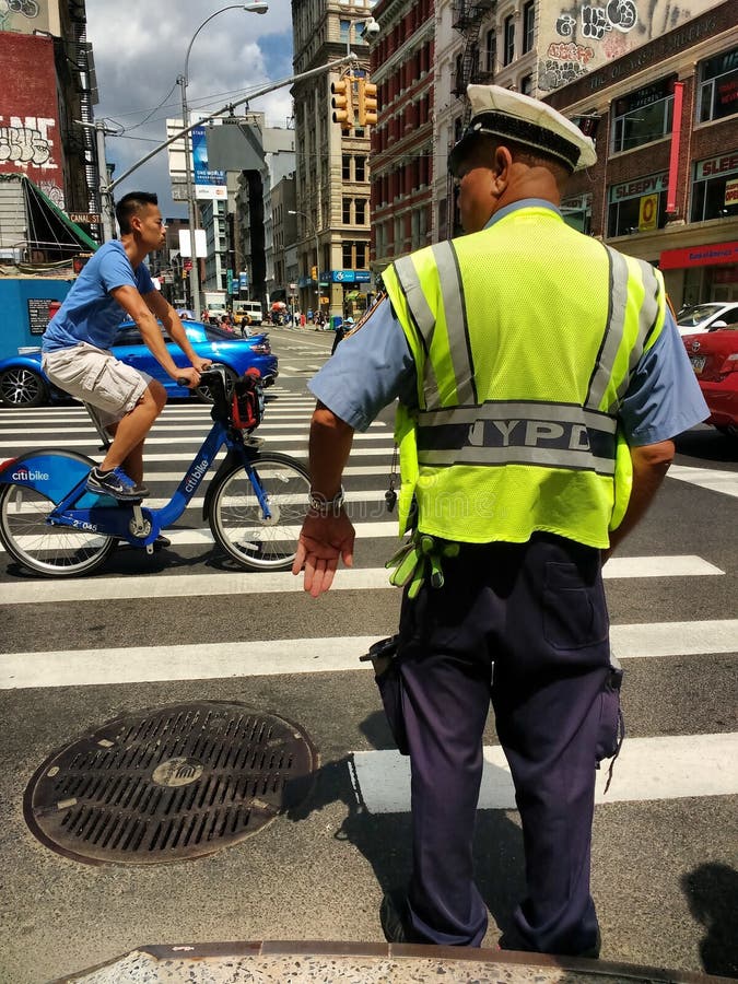 An NYPD officer wearing a high-visibility yellow reflective safety vest stands on the corner of Canal Street and Broadway, signaling to cars, pedestrians, and cyclists. A young man rides past on a Citi Bike. An NYPD officer wearing a high-visibility yellow reflective safety vest stands on the corner of Canal Street and Broadway, signaling to cars, pedestrians, and cyclists. A young man rides past on a Citi Bike.