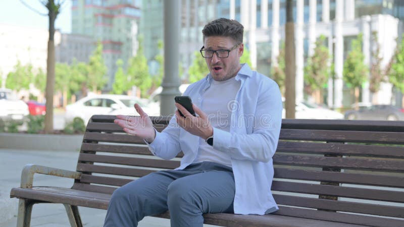När man reagerar på förlust på smartphone när man sitter utomhus på bänk royaltyfri fotografi