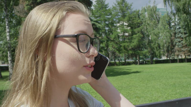 Nära lugn, blond, ung kvinna i glasögon som har ett viktigt telefonsamtal som sitter på parken framför stora