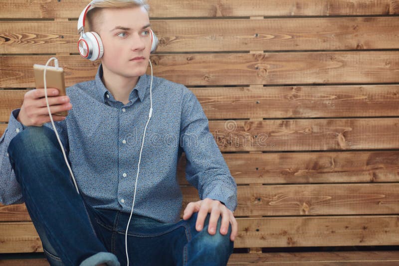 Närbild av den hållande mobiltelefonen för ung scandinavian man och lyssnande musik med leende, medan sitta på trägolvet royaltyfri bild Närbild av den hållande mobiltelefonen för ung scandinavian man och lyssnande musik med leende, medan sitta på trägolvet royaltyfri bild