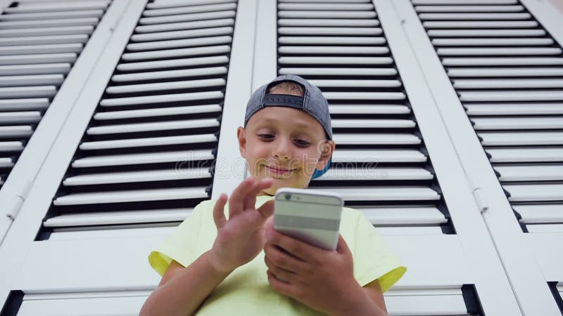 Närbild av pysen som använder mobiltelefonen, medan att spela spelar Stående av den bärande T-tröja för gullig pojke som skriver stock video