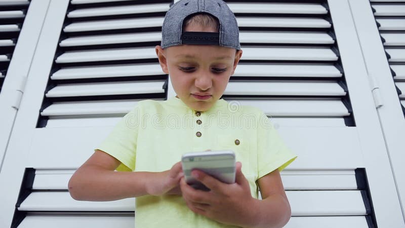 Närbild av pysen som använder mobiltelefonen, medan att spela spelar Ståenden av använda för T-tröja för härlig pojke bärande är arkivfilmer