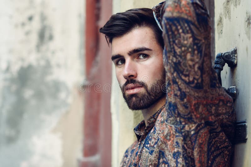 Close-up portrait of young bearded man, model of fashion, in urban background wearing casual clothes. Guy with beard and modern hairstyle in the street. Close-up portrait of young bearded man, model of fashion, in urban background wearing casual clothes. Guy with beard and modern hairstyle in the street.