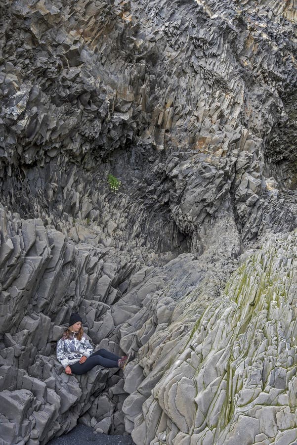 O adolescente da menina está sentando-se confortavelmente em penhascos do basalto no fundo da montanha de Reynisfjall Praia de Re foto de stock O adolescente da menina está sentando-se confortavelmente em penhascos do basalto no fundo da montanha de Reynisfjall Praia de Re foto de stock