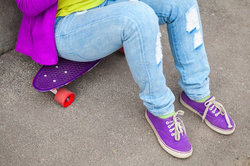 O adolescente nas calças de brim e nos gumshoes senta-se em um skate imagem de stock O adolescente nas calças de brim e nos gumshoes senta-se em um skate imagem de stock