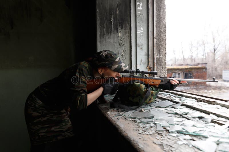 O atirador furtivo da menina olha com o espa?o O guerreiro da menina dirige seu rifle atrav?s da janela quebrada de uma constru?? imagem de stock O atirador furtivo da menina olha com o espa?o O guerreiro da menina dirige seu rifle atrav?s da janela quebrada de uma constru?? imagem de stock
