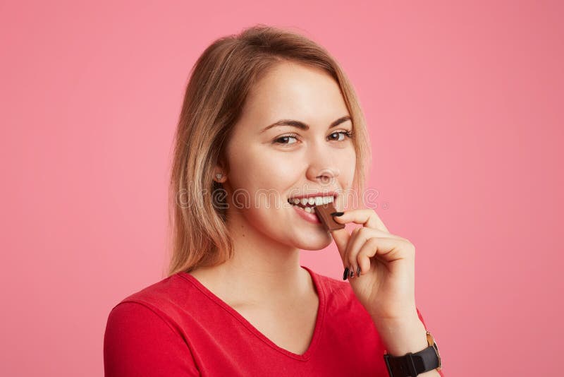 O chocolate doce delicioso das mordidas fêmeas satisfeitas satisfeitos, aprecia seu gosto, tem a expressão feliz e o olhar agradá fotografia de stock royalty free O chocolate doce delicioso das mordidas fêmeas satisfeitas satisfeitos, aprecia seu gosto, tem a expressão feliz e o olhar agradá fotografia de stock royalty free