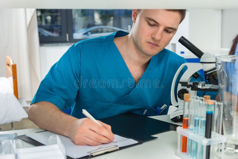 O cientista masculino novo no uniforme está escrevendo para baixo notas de seu res fotografia de stock O cientista masculino novo no uniforme está escrevendo para baixo notas de seu res fotografia de stock