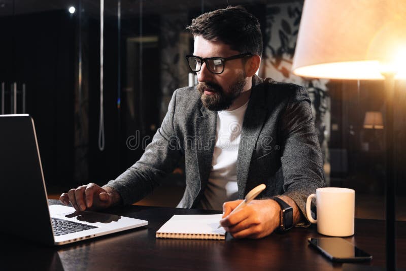O colega de trabalho farpado senta-se pela tabela de madeira com lâmpada e utilização do caderno contemporâneo Pena da posse do h imagem de stock O colega de trabalho farpado senta-se pela tabela de madeira com lâmpada e utilização do caderno contemporâneo Pena da posse do h imagem de stock
