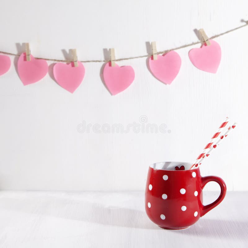 O copo vermelho em um ponto branco está em uma tabela de madeira branca Cair de papel dos corações na parede fotos de stock royalty free O copo vermelho em um ponto branco está em uma tabela de madeira branca Cair de papel dos corações na parede fotos de stock royalty free
