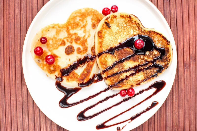 Heart shaped pancakes with chocolate sauce and red cranberries on white porcelain plate. Celebration dessert. Indoors closeup. Heart shaped pancakes with chocolate sauce and red cranberries on white porcelain plate. Celebration dessert. Indoors closeup.