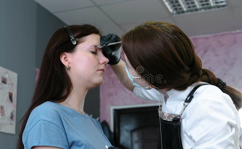 O Cosmetologist põe a pintura sobre as sobrancelhas do ` s do cliente com escova Vista lateral Correção da sobrancelha imagem de stock royalty free O Cosmetologist põe a pintura sobre as sobrancelhas do ` s do cliente com escova Vista lateral Correção da sobrancelha imagem de stock royalty free