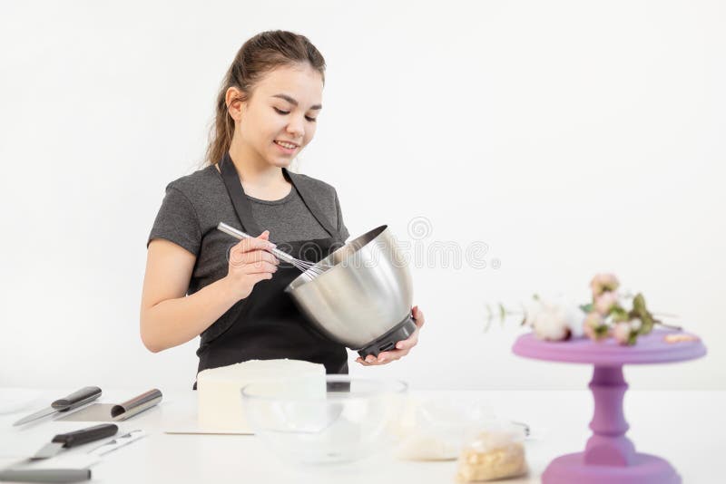 O cozinheiro chefe de pastelaria moreno atrativo da menina no avental chicoteia o bolo de creme em uma bacia do metal imagens de stock O cozinheiro chefe de pastelaria moreno atrativo da menina no avental chicoteia o bolo de creme em uma bacia do metal imagens de stock
