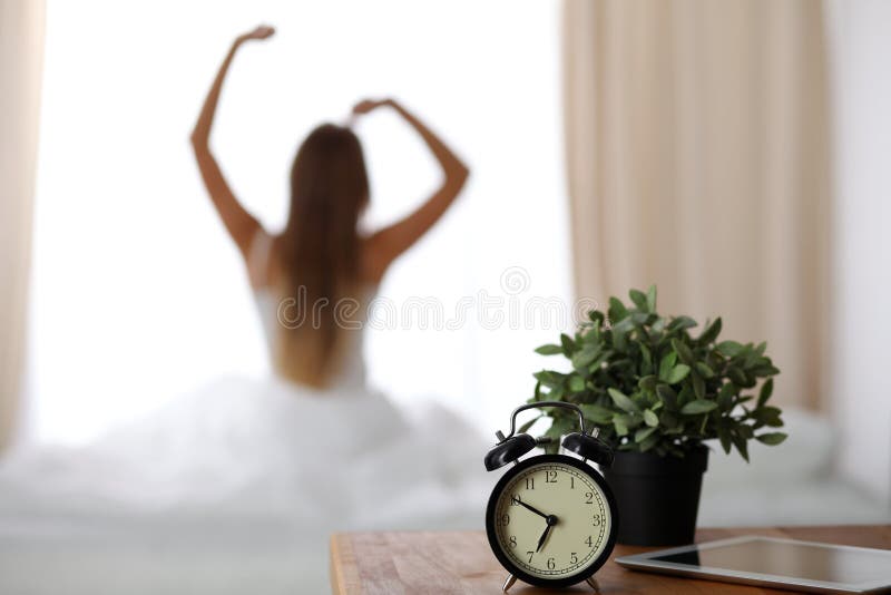 O despertador que está na tabela de cabeceira tem já soou o amanhecer para acordar a mulher está esticando na cama no fundo fotografia de stock O despertador que está na tabela de cabeceira tem já soou o amanhecer para acordar a mulher está esticando na cama no fundo fotografia de stock