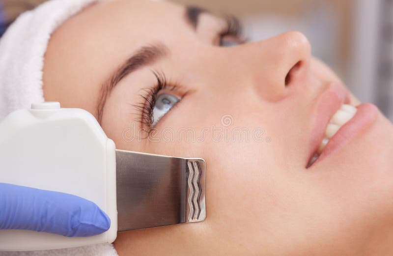 O doutor-cosmetologist faz o procedimento de limpeza do ultrassom da pele facial de um bonito, jovem mulher imagem de stock royalty free O doutor-cosmetologist faz o procedimento de limpeza do ultrassom da pele facial de um bonito, jovem mulher imagem de stock royalty free