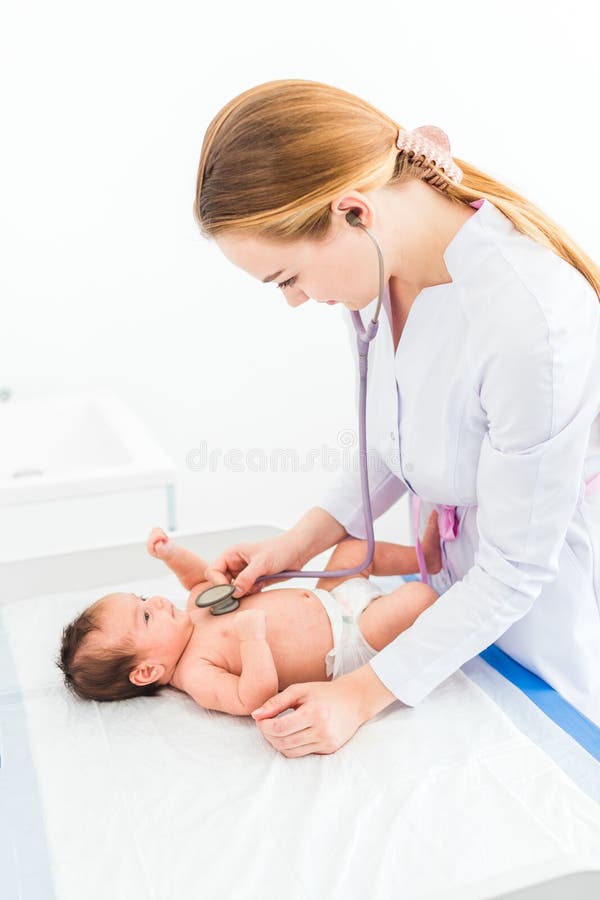 O doutor louro fêmea novo bonito do pediatra examina o bebê com estetoscópio que verifica o batimento cardíaco imagem de stock royalty free O doutor louro fêmea novo bonito do pediatra examina o bebê com estetoscópio que verifica o batimento cardíaco imagem de stock royalty free